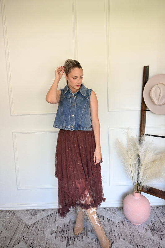 Woman in a denim vest and brown skirt standing in a room with a white wall and patterned floor.