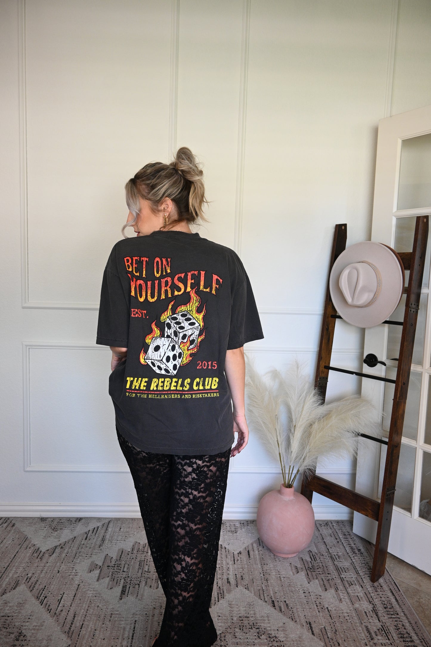 Person wearing a black graphic tee with text and graphics, standing in front of a wall.