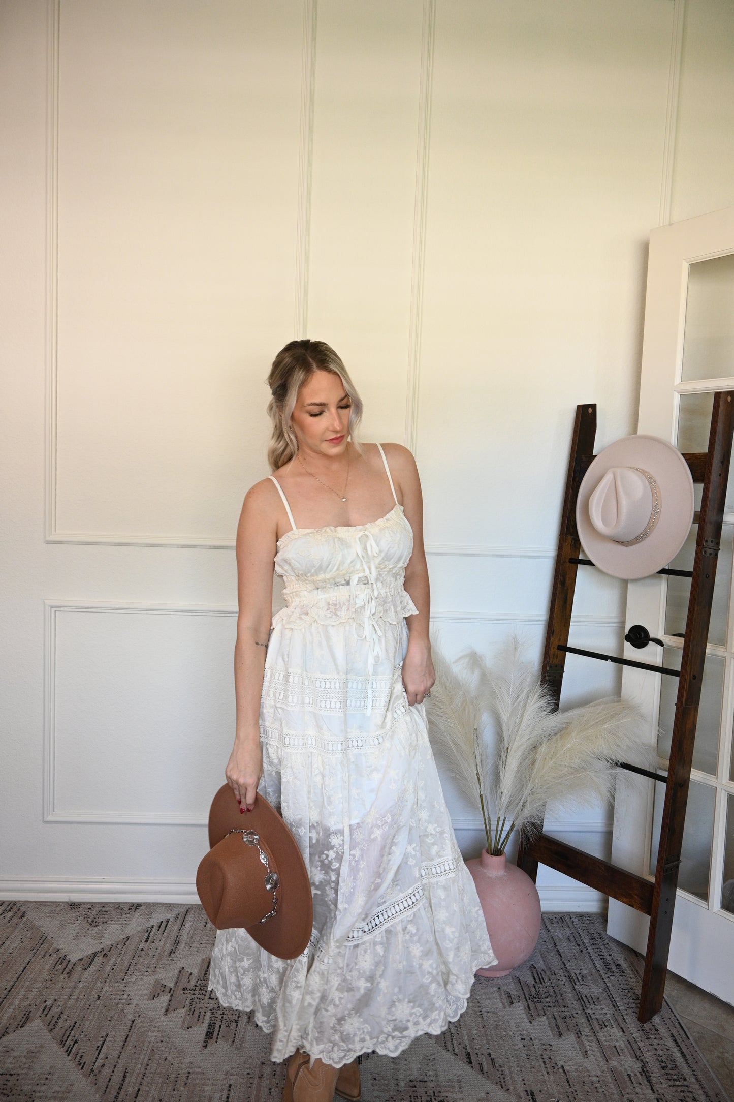 Woman in a ivory top and skirt set holding a brown hat in a room with a wooden floor and white walls.