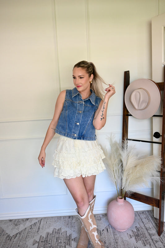 Woman in denim vest and white skirt standing in a room with a hat rack and vase.