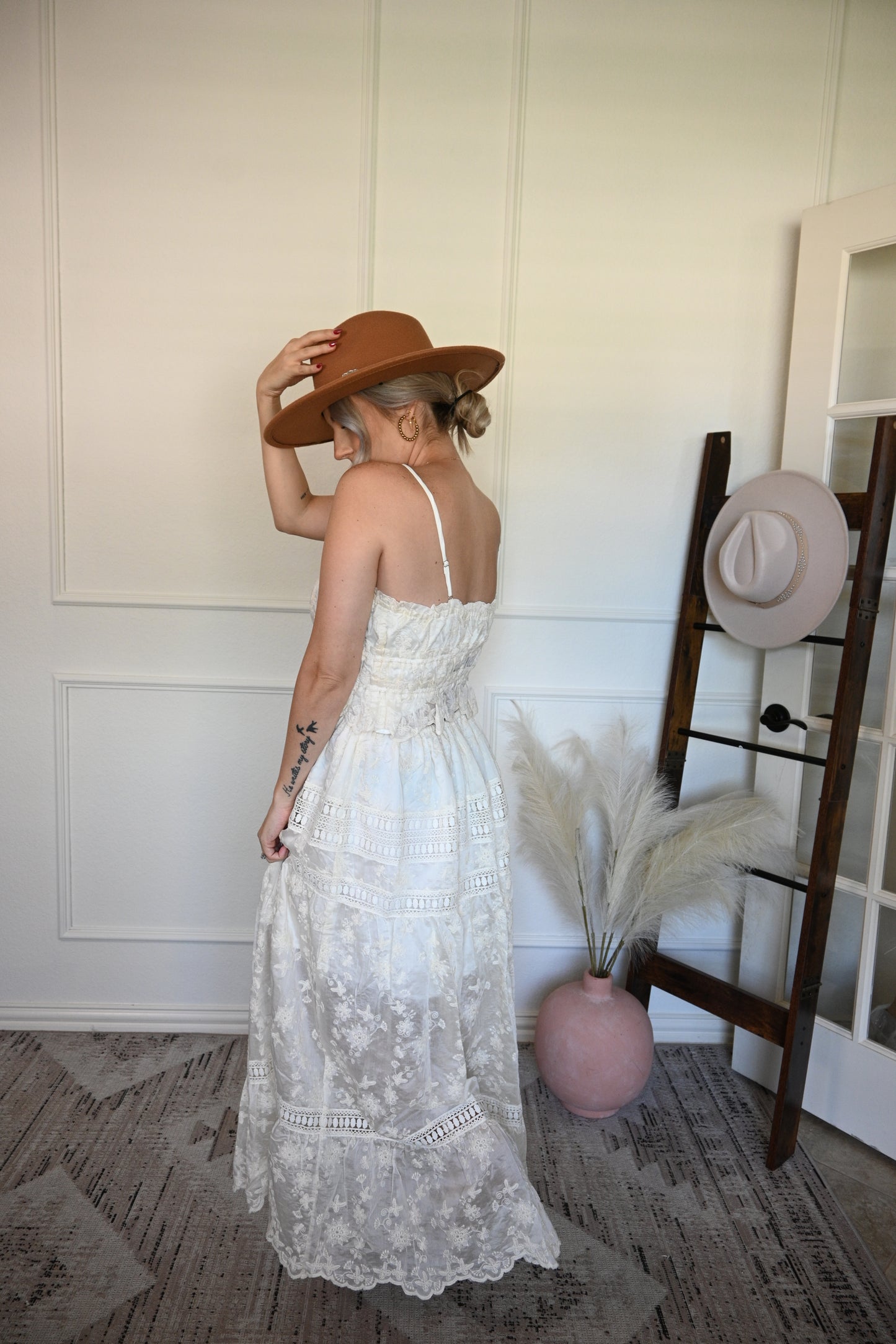 Woman in a ivory top and lace skirt set and brown hat standing next to a hat rack with hats on it.