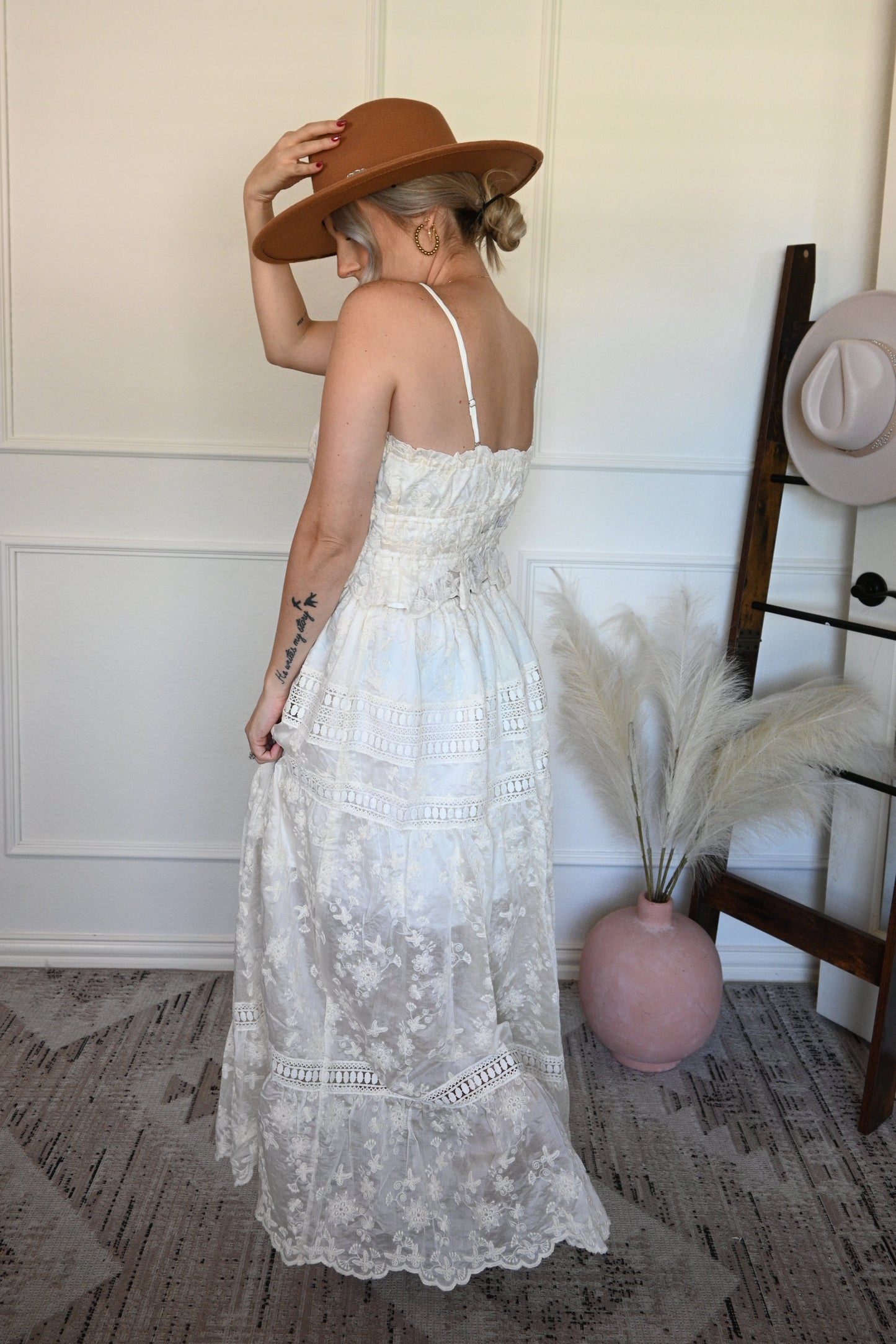 Woman in a ivory top and lace skirt set and brown hat standing next to a hat rack with hats on it.