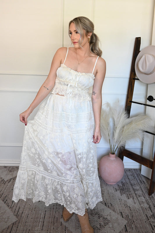 Woman holding one side of the top and lace skirt set in a room with a wooden floor and white walls.