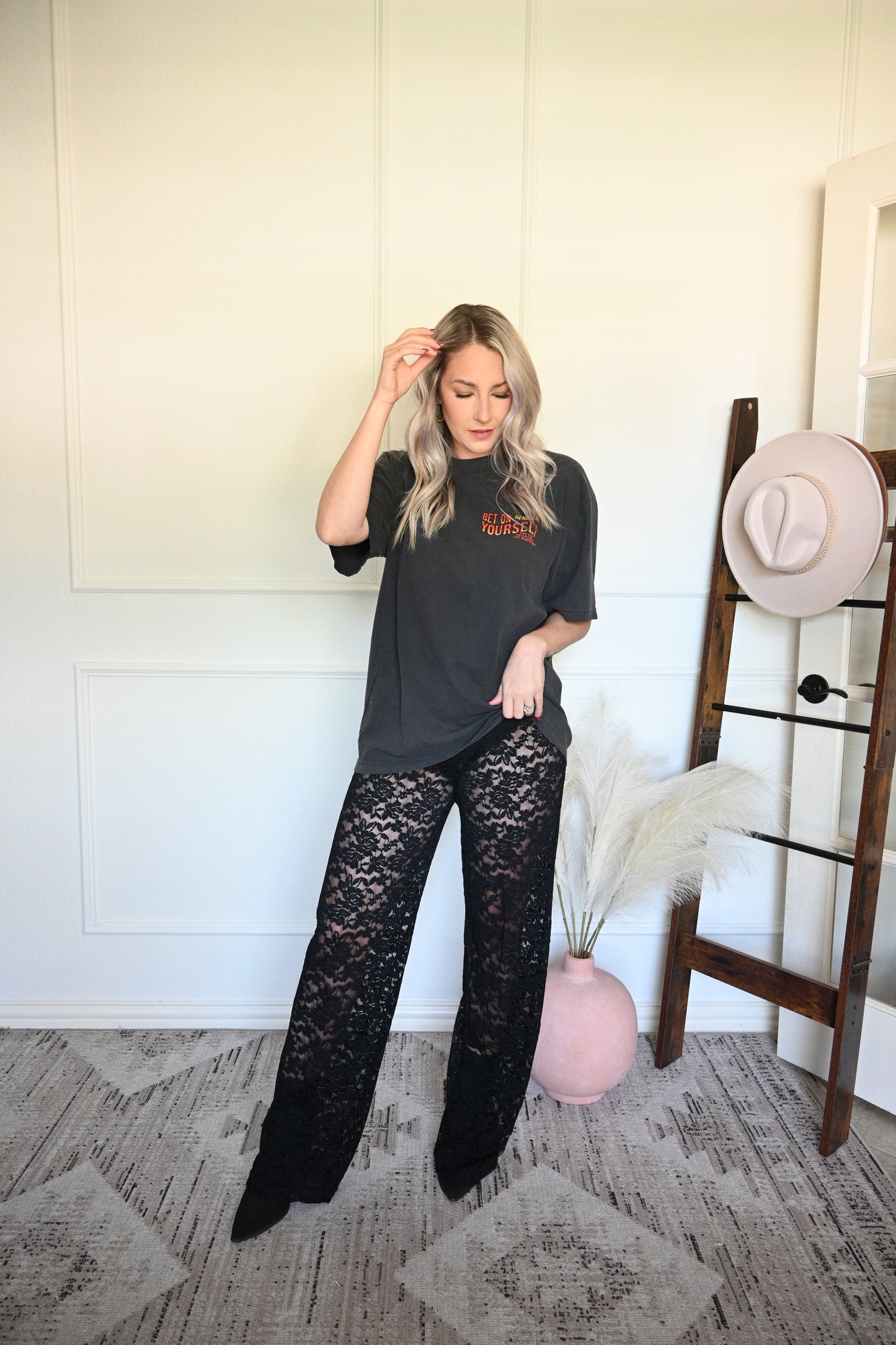 Woman in black lace pants and black oversized graphic tee with a white wall and patterned rug.