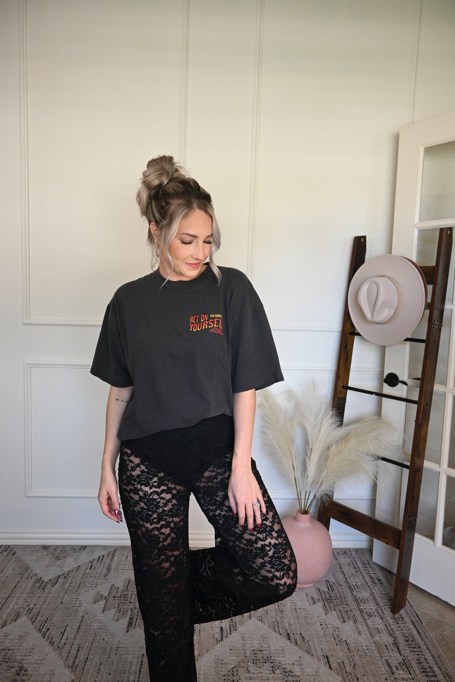 Person wearing a black graphic tee tucked in to black sheer lace pants standing on a decorative rug