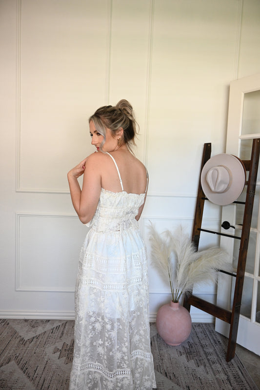 Woman in a ivory lace tank top and skirt set tanding next to a hat on a rack against a white wall.