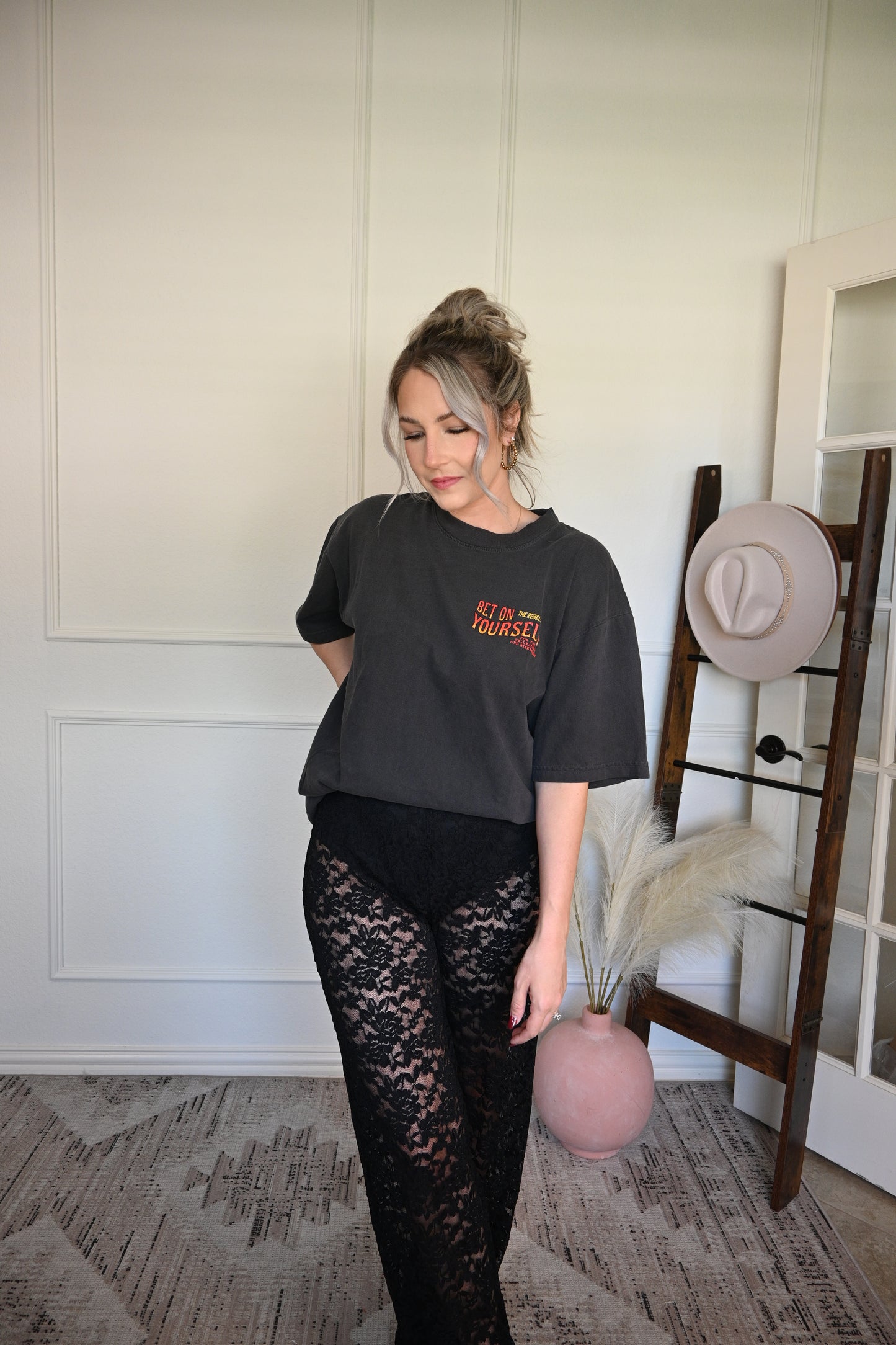 Person wearing an oversized graphic tee with text and lace pants standing in a room with a mirror and hat.