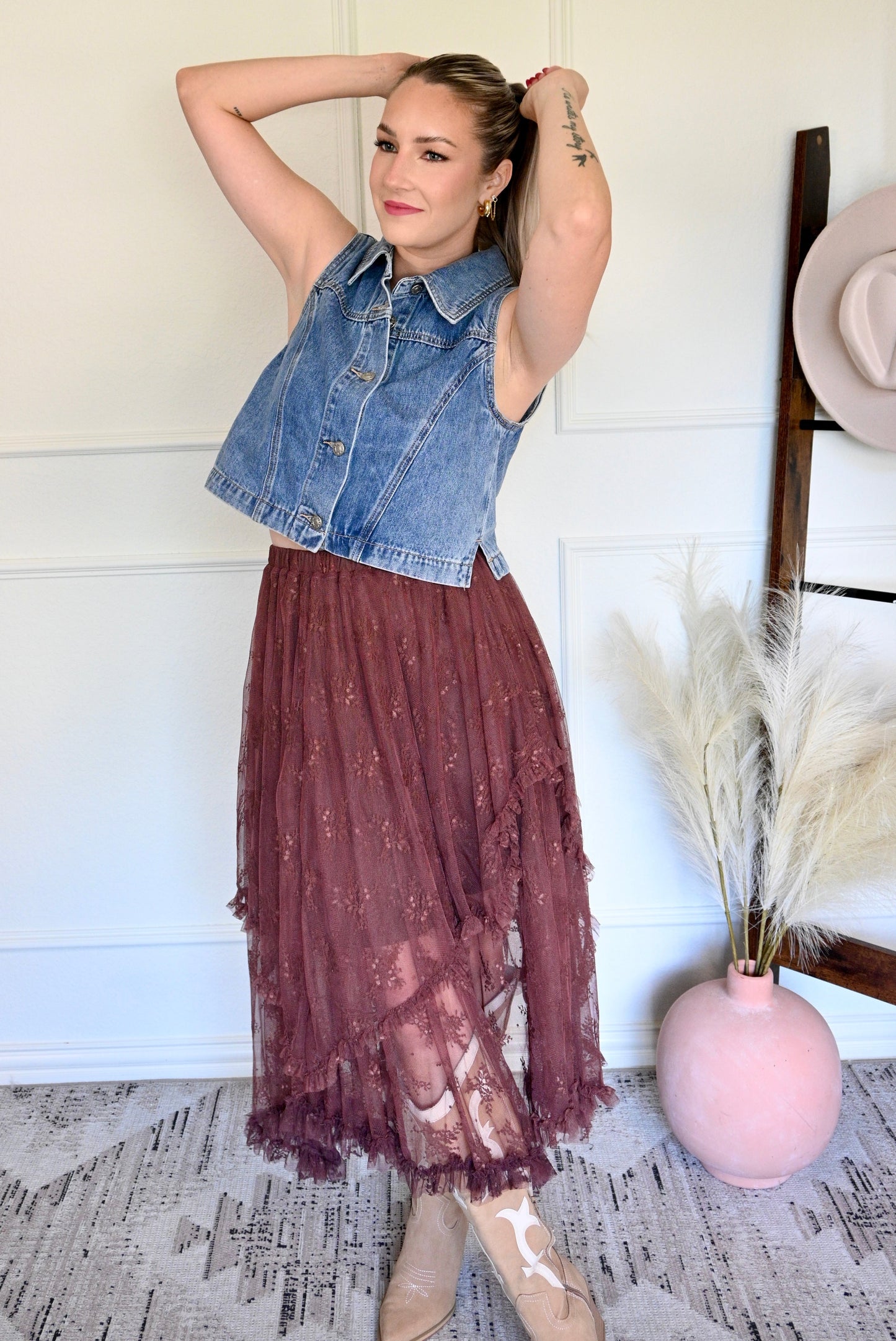 Woman wearing a denim vest over a lace dress in a room with a white wall and decorative elements.