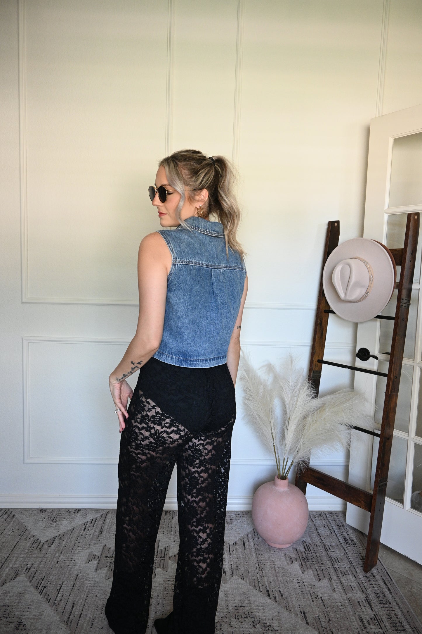 Woman in denim vest and black sheer lace pants standing next to a hat rack with hats on a white wall.