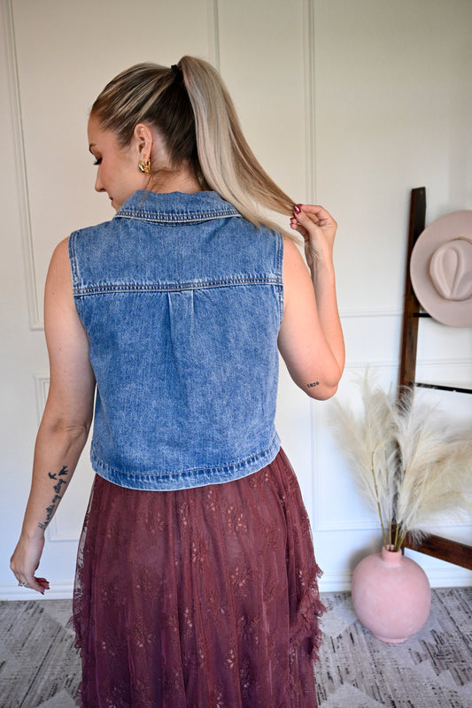 Woman wearing a denim vest with a brown skirt, standing in a room with a white wall and a pink vase.