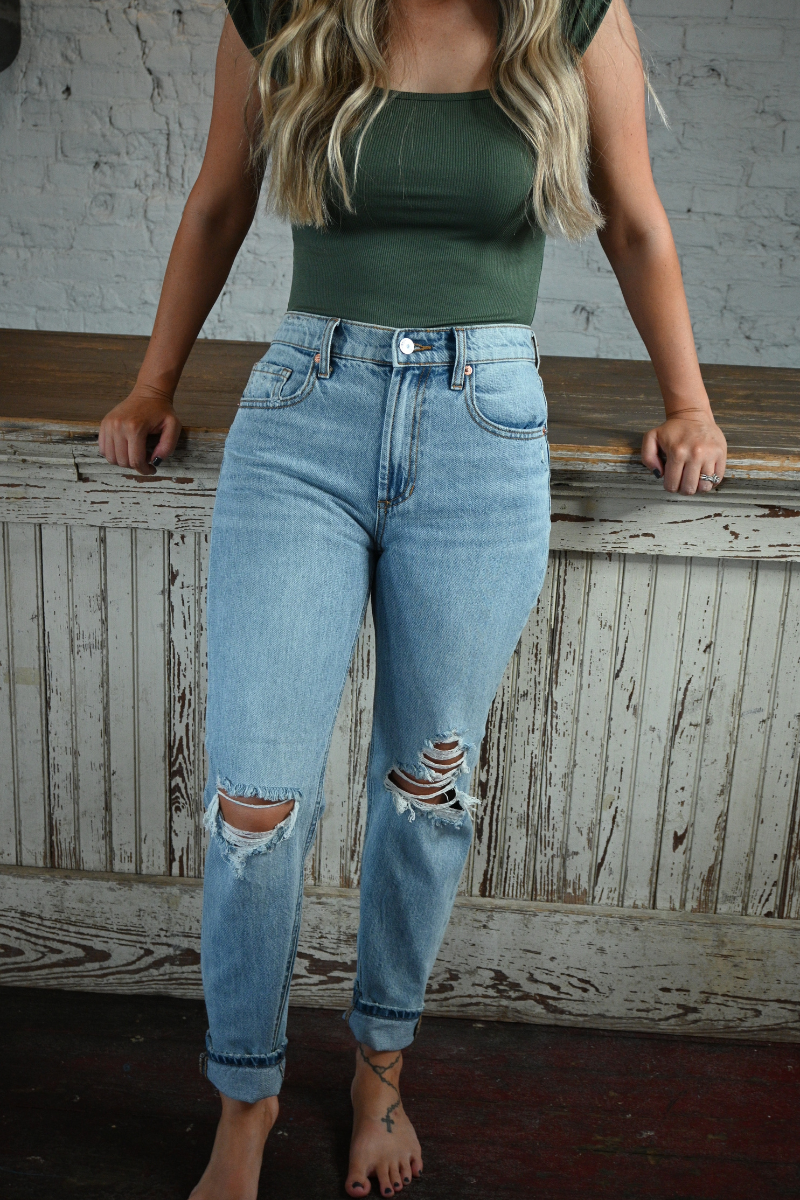 Woman wearing high rise light wash skinny jeans and a green tight fit shirt leaning on a distressed wood bar.