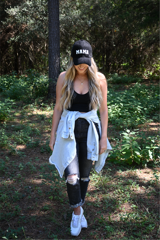A woman standing looking down wearing a black corduroy mama hat with a black ribbed cropped cami and dark grey distressed skinny jeans with a distressed denim jacket around her waist.