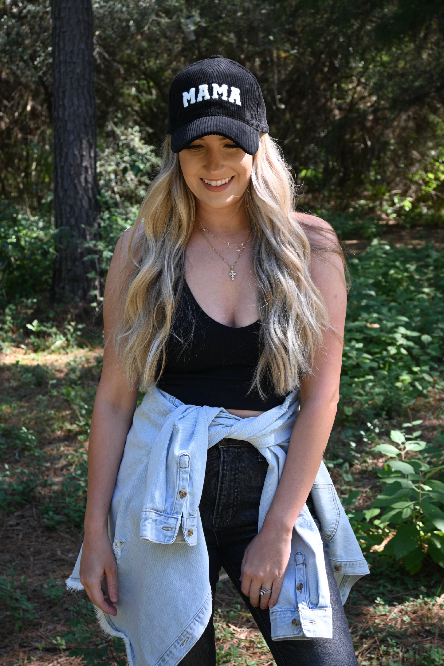 A woman is smiling looking down wearing a black corduroy mama hat and a black ribbed cropped cami with dark grey distressed skinny jeans and a distressed denim jacket around her waist.