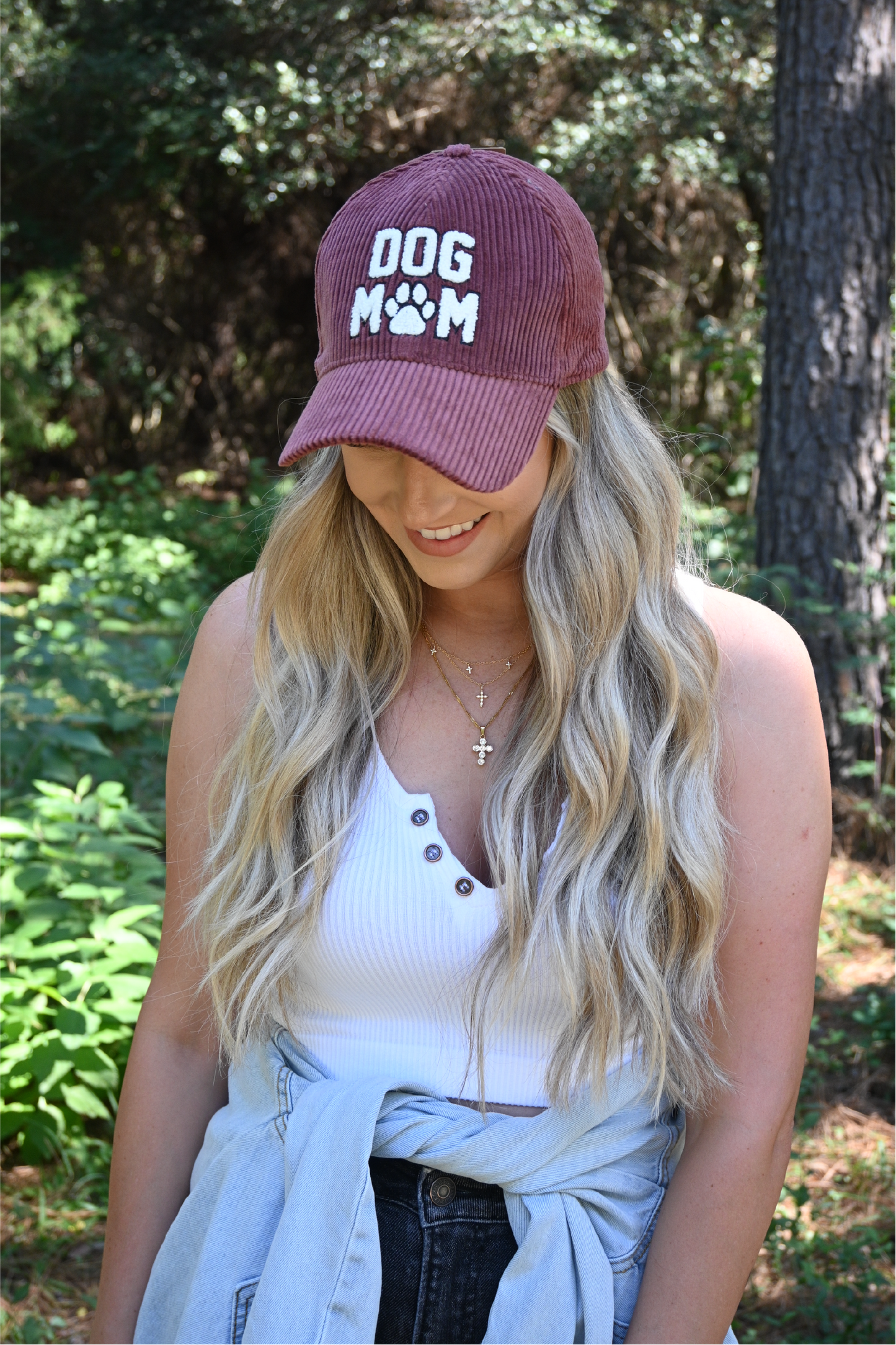A woman is smiling looking down wearing a burgundy corduroy dog mom hat with a white ribbed cropped cami and dark grey distressed skinny jeans with a distressed denim jacket around her waist.