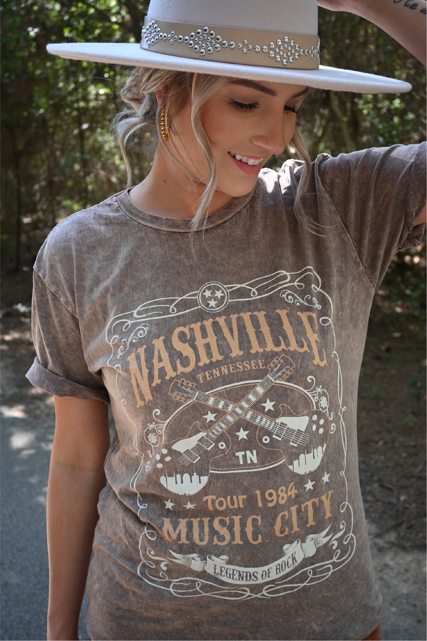 A close up shot of a woman smiling looking down wearing the nashville graphic tee and the ivory faux leather band fedora hat.
