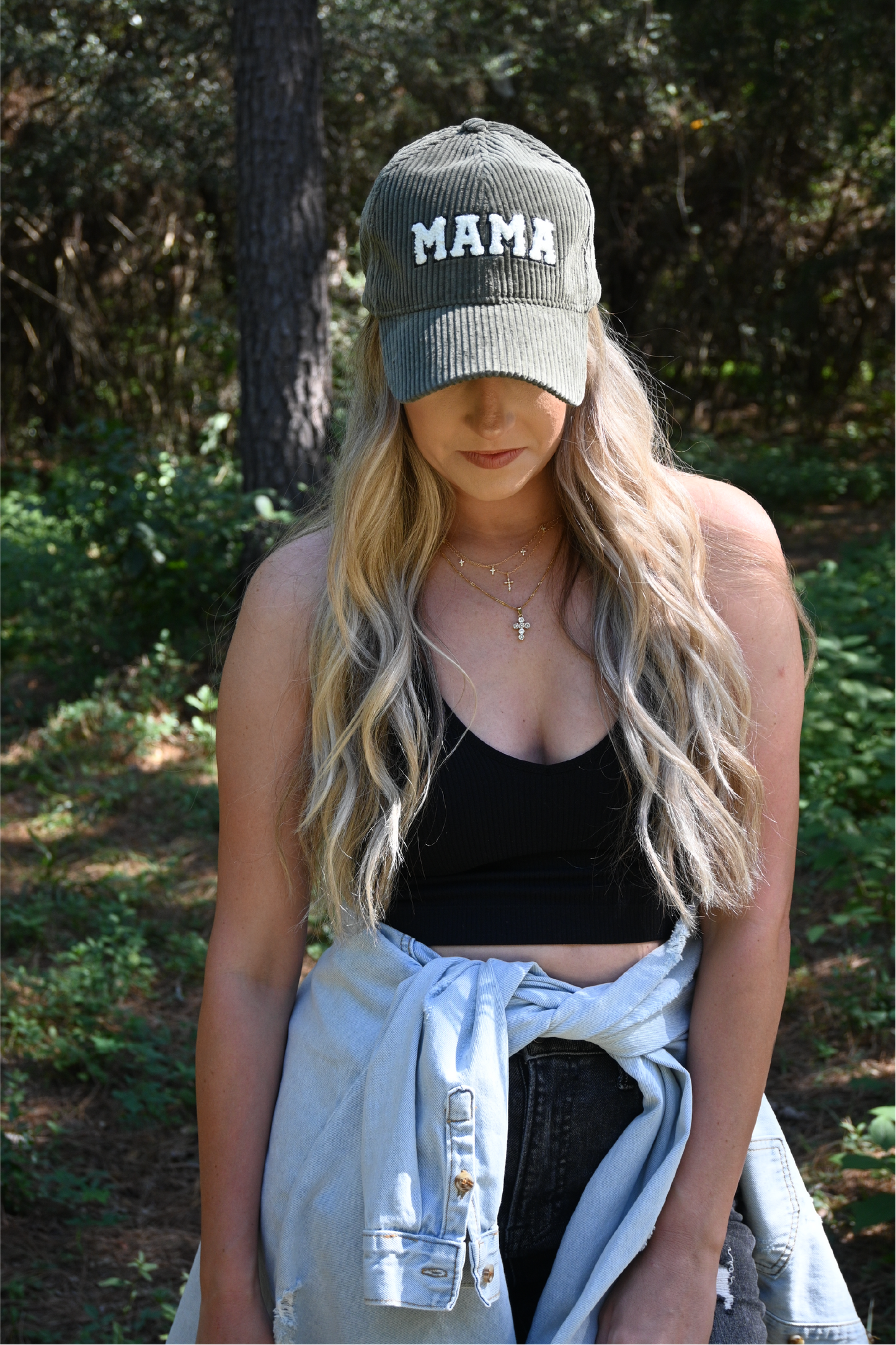 A woman is looking down wearing a olive corduroy mama hat with a black ribbed cropped cami and dark grey distressed skinny jeans with a distressed denim jacket around her waist.