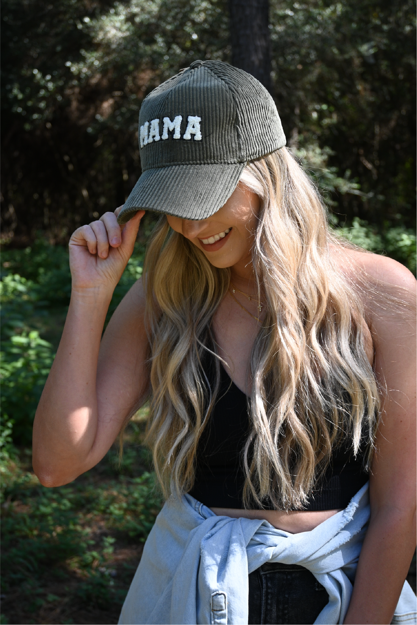 A woman is is smiling looking down with one hand on the brim of an olive corduroy mama hat. She is also wearring a black ribbed cropped cami and dark grey distressed skinny jeans and a distressed denim jacket around her waist.