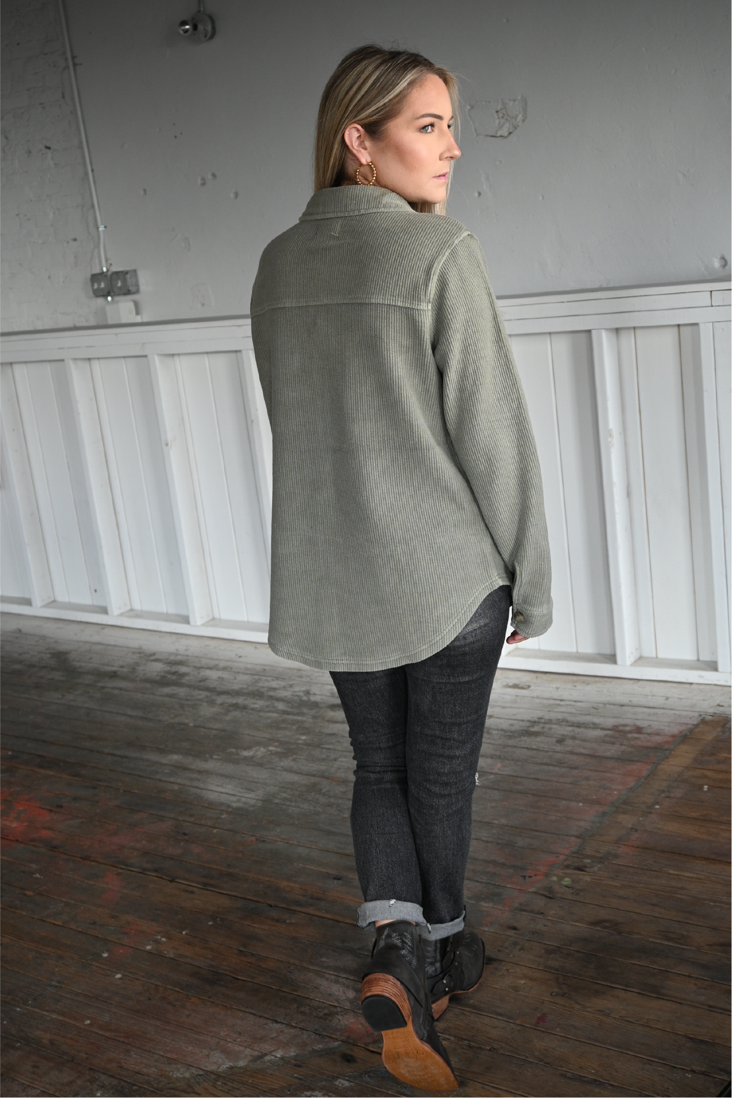A woman has her back turned to the camera looking over her shoulder wearing the olive shacket paired with the dark grey skinny jeans cuffed at the bottom with the pierre dumas buckle ankle boots.