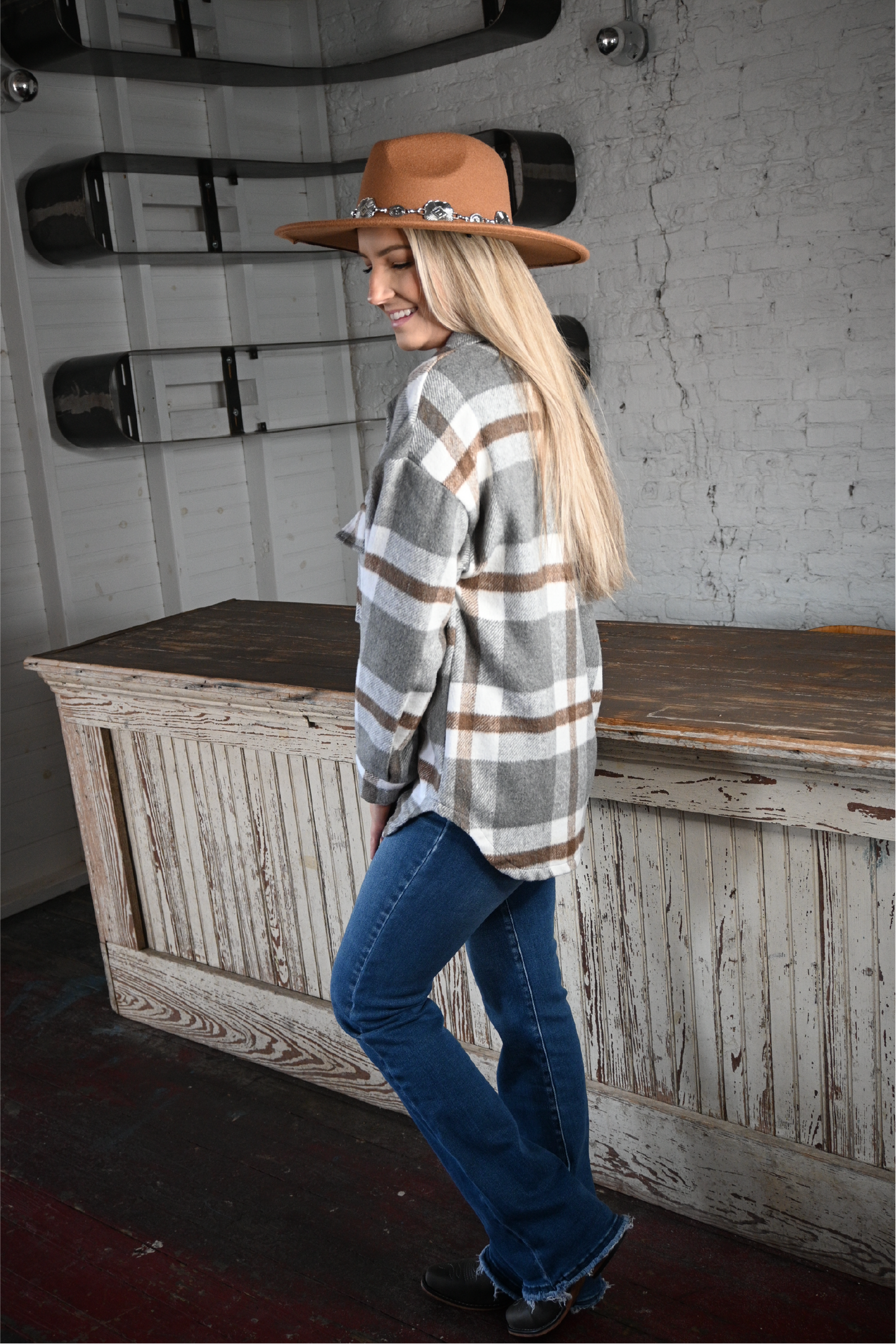 A woman is turned to the side smiling wearing the tan silver strap flat brim fedora hat and plaid flannel shacket paired with the high rise bootcut kancan jeans and pierre dumas buckle ankle boots.