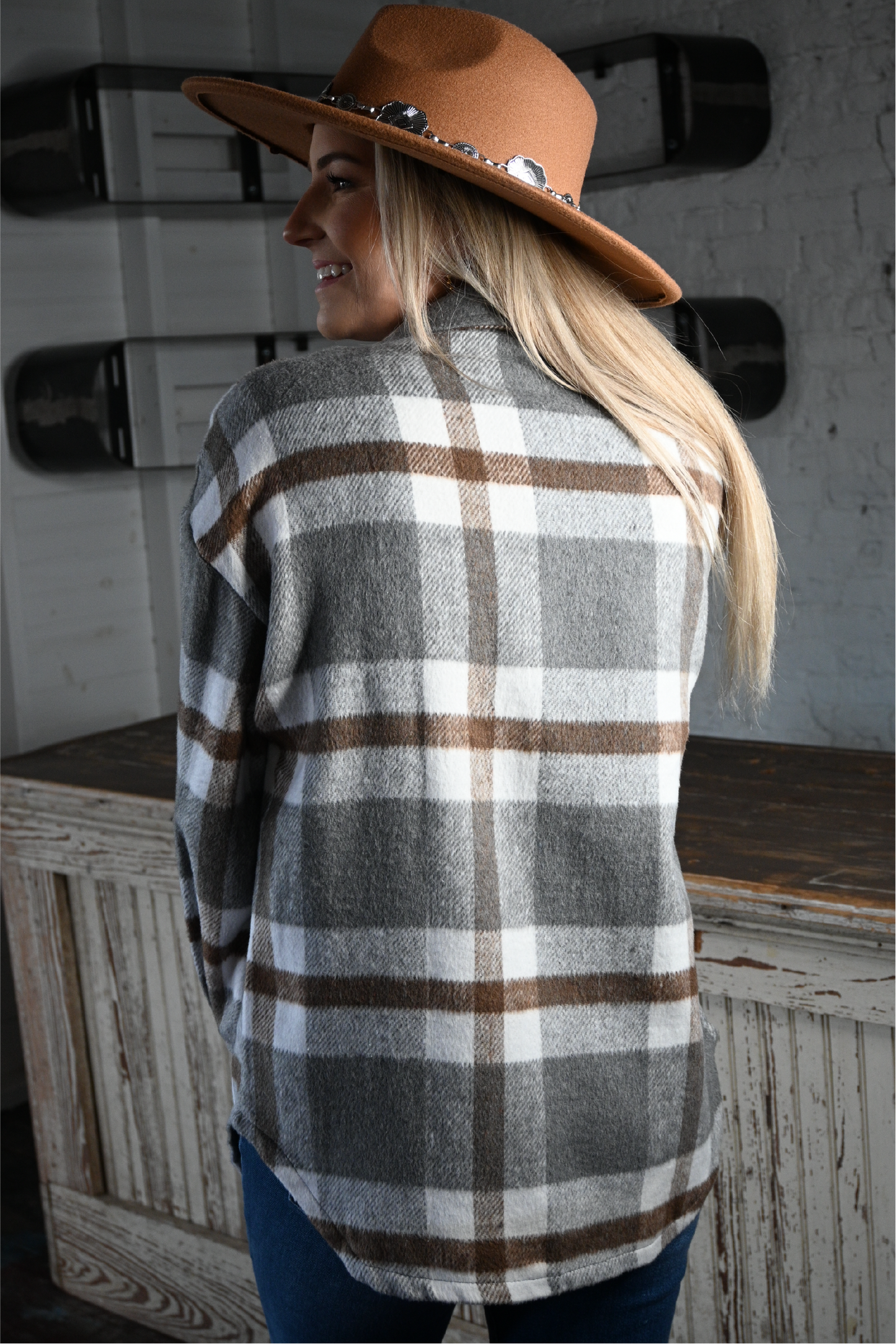 A woman has her back turned towards the camera looking over her shoulder smiling wearing the tan silver strap flat brim fedora hat and plaid flannel shacket.