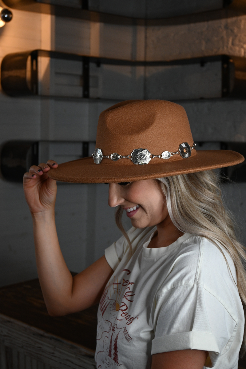 Side view of a woman smiling and holding the top of her tan fedora hat.
