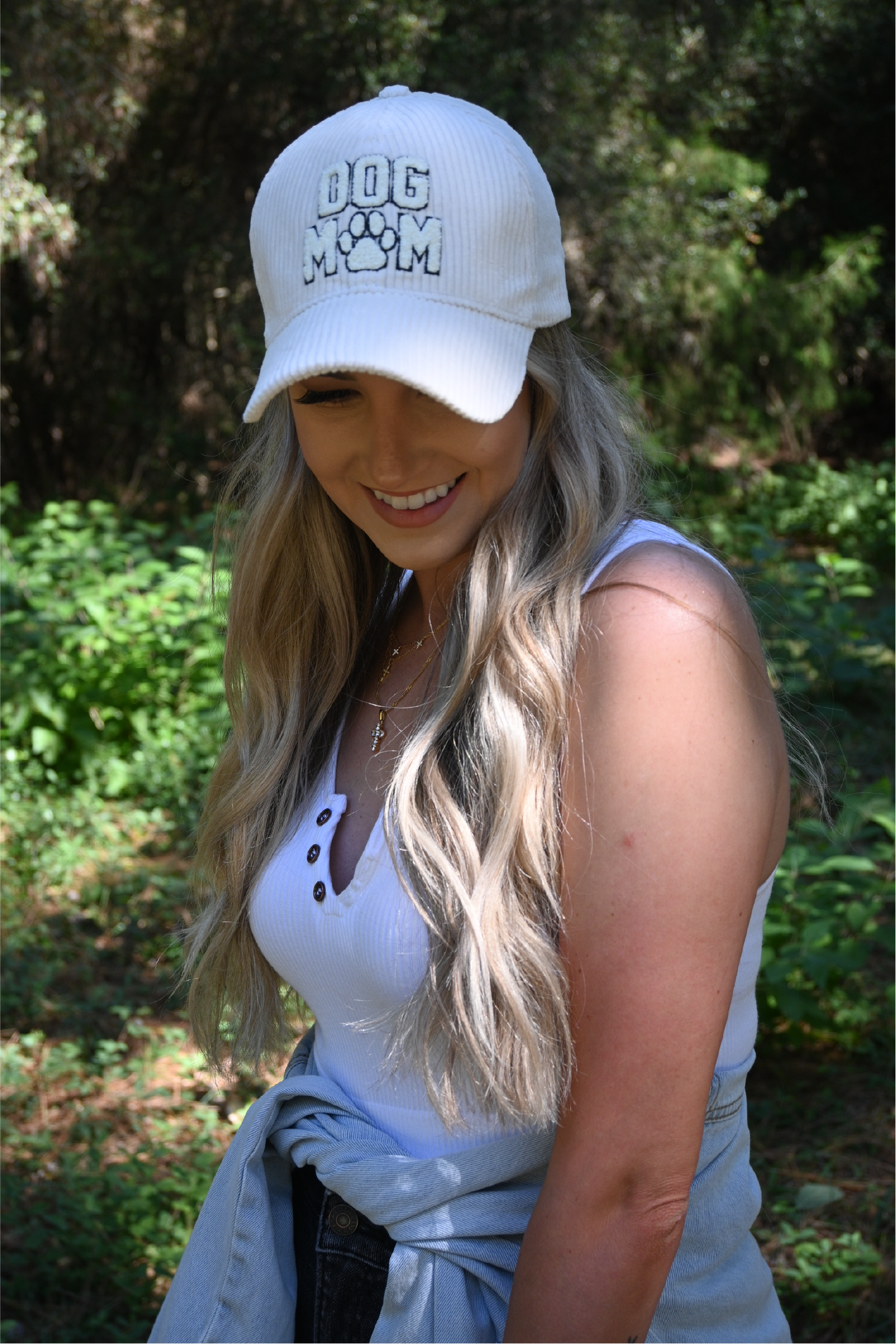 A woman smiling looking down wearing a white corduroy dog mom hat and a white ribbed cropped cami with a distressed denim jacket around her waist.