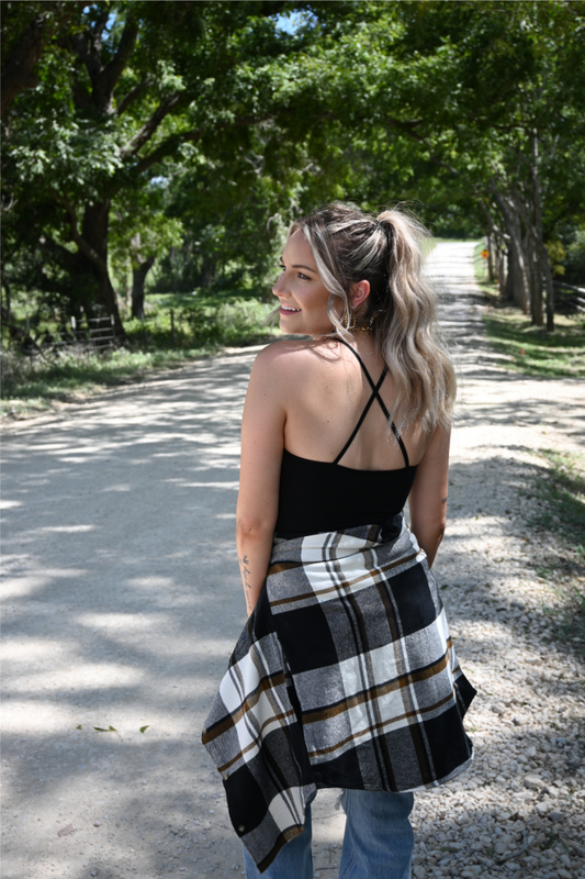 A women has her back to the camera looking over her shoulder wearing the black ribbed cropped cami paired with the 90's skinny high rise jeans and the black brown plaid flannel button down tied around her waist.