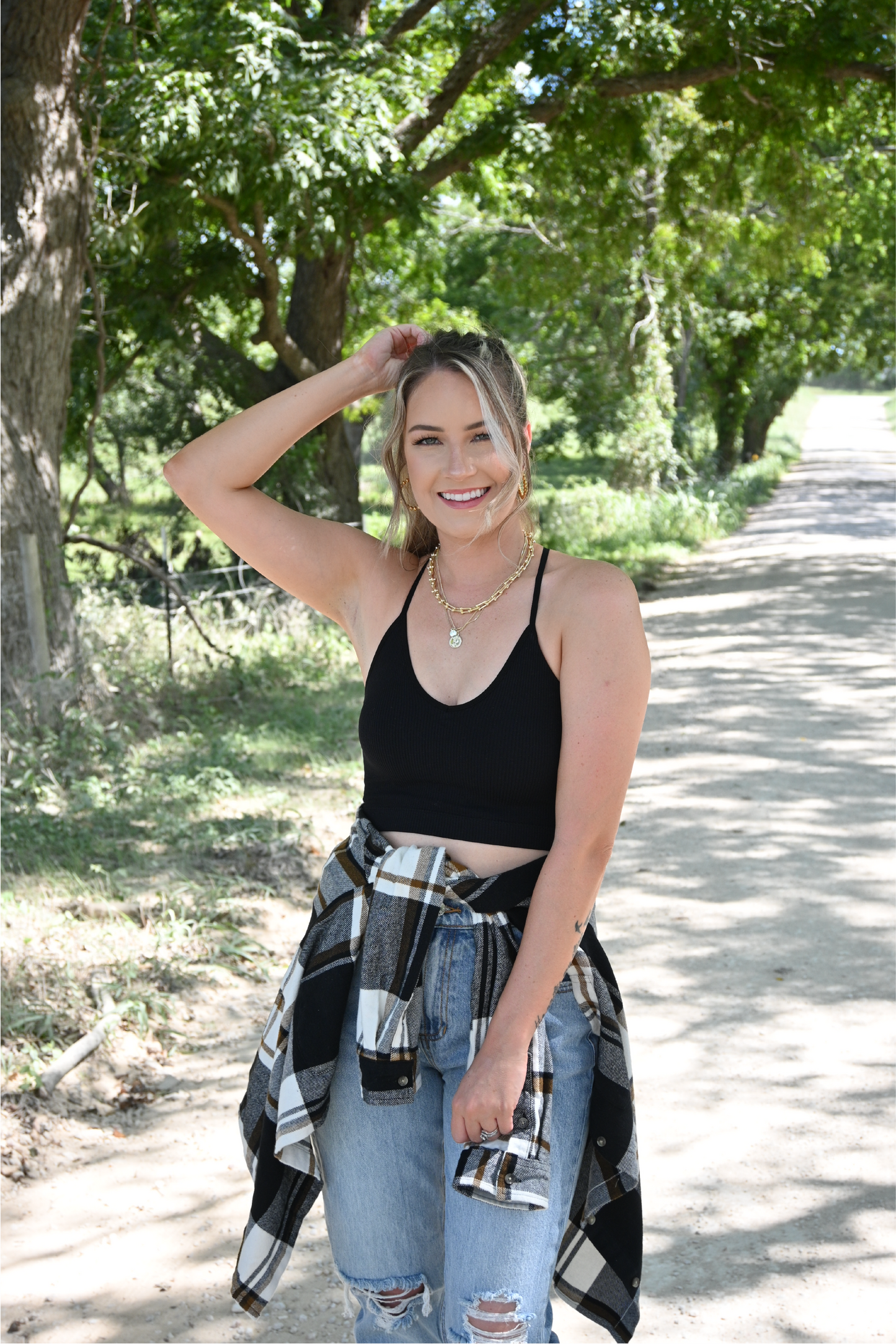 A women is smiling at the camera on a dirt road with her right hand on her head and the other by her side. She is wearing the black ribbed cropped cami paired with the 90's skinny high rise jeans and black brown plaid flannel button down tied around her waist. She is also wearing the gold chain necklace and the gold coin necklace.