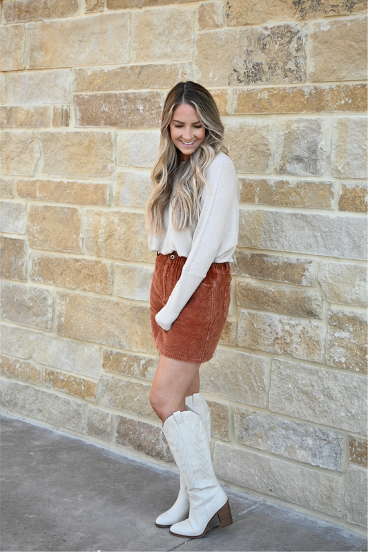 A woman is turned to the side looking down smiling while her hand is at her side wearing the oatmeal crew neck sweater tucked into the corduroy paper bag skirt and ivory knee high MIA boots