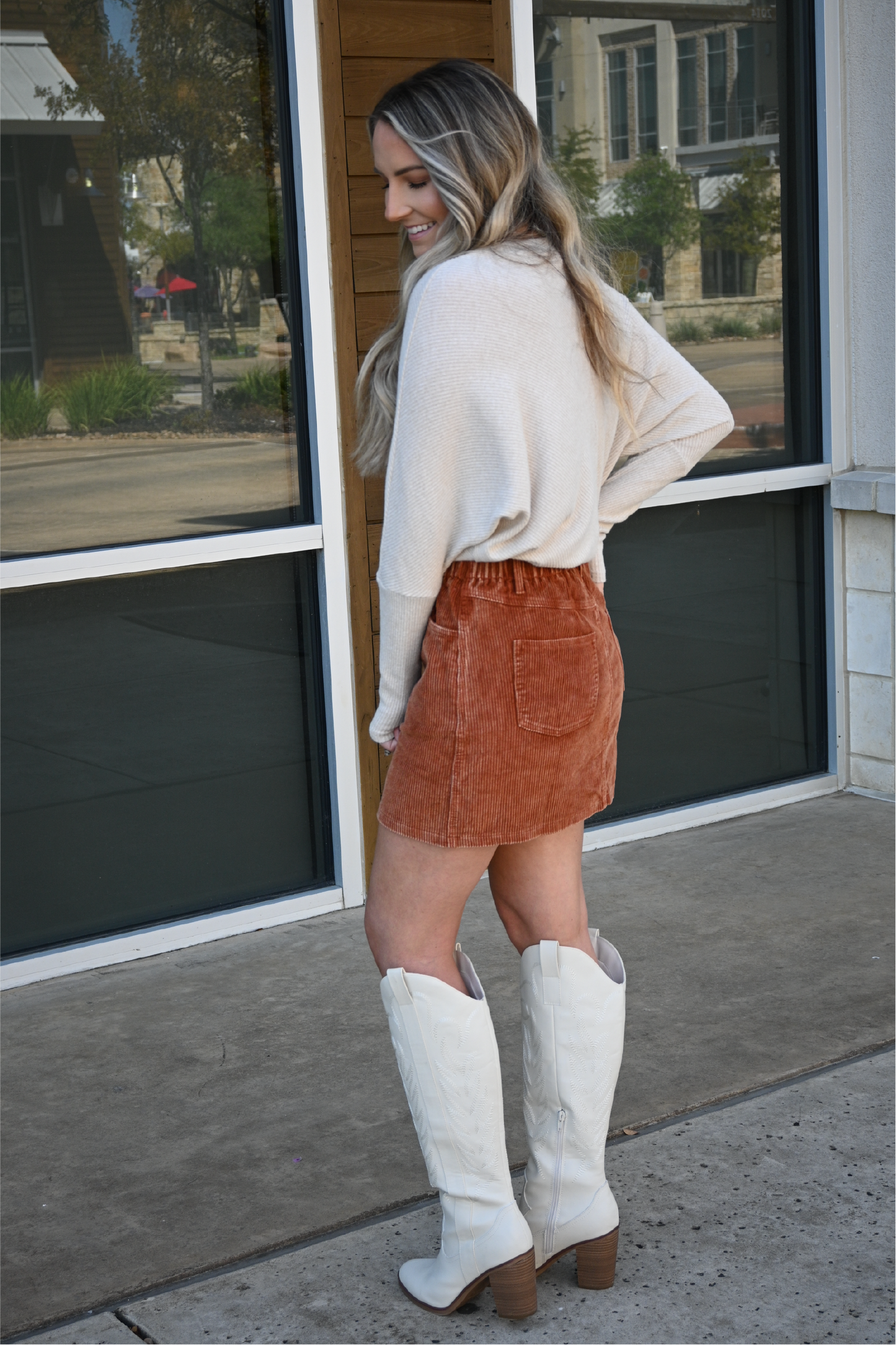 A woman has her back turned to the camera while looking over her shoulder smiling wearing the corduroy paper bag skirt with the oatmeal crew neck sweater tucked in and wearing the ivory knee high MIA boots.