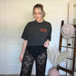Person wearing a black graphic tee with text and lace pants, standing next to a chair with hats on it.