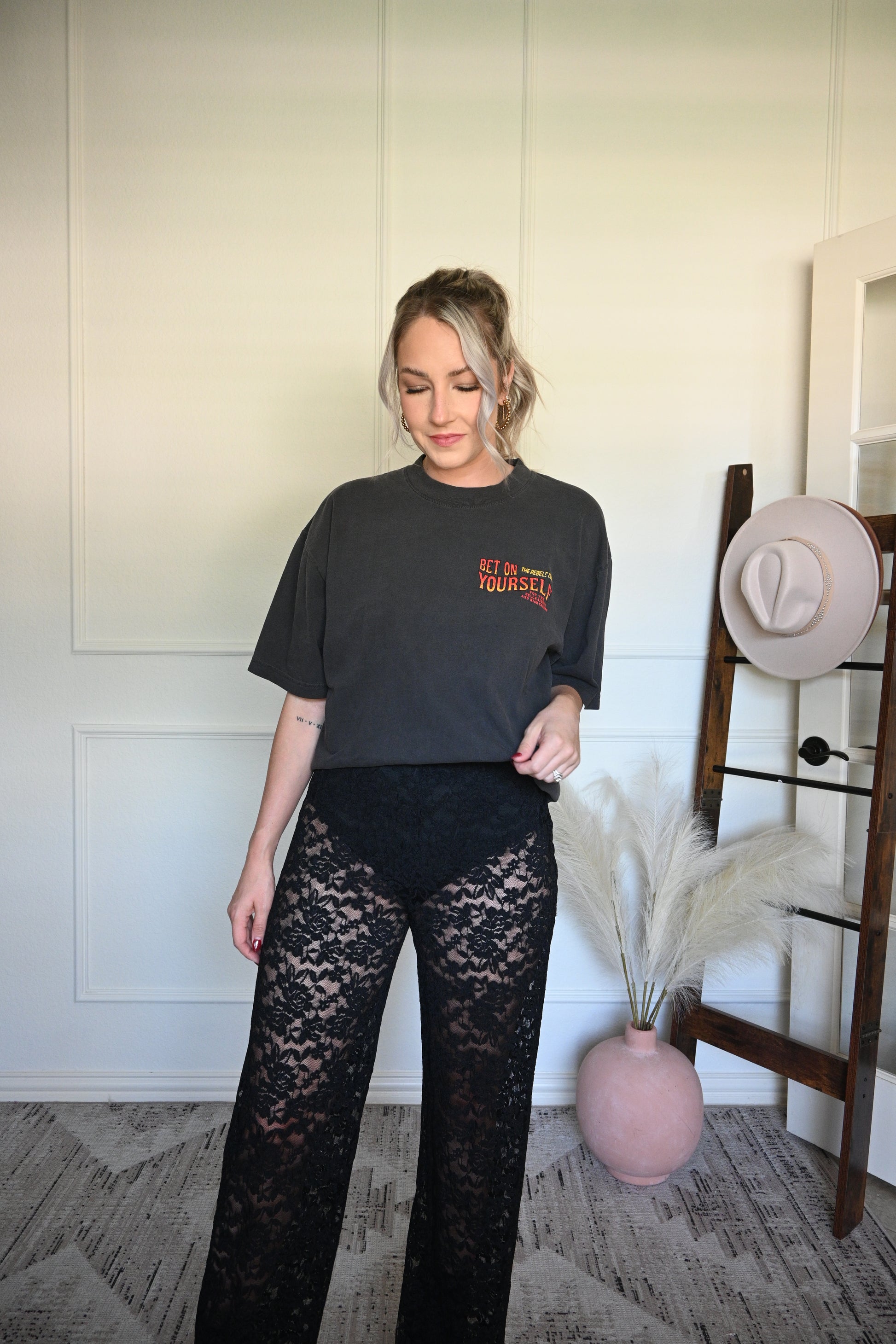Person wearing a black graphic tee with text and lace pants, standing next to a chair with hats on it.