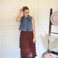 Woman in a denim vest and brown skirt standing in a room with a white wall and patterned floor.