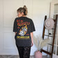 Person wearing a black graphic tee with text and graphics, standing in front of a wall.