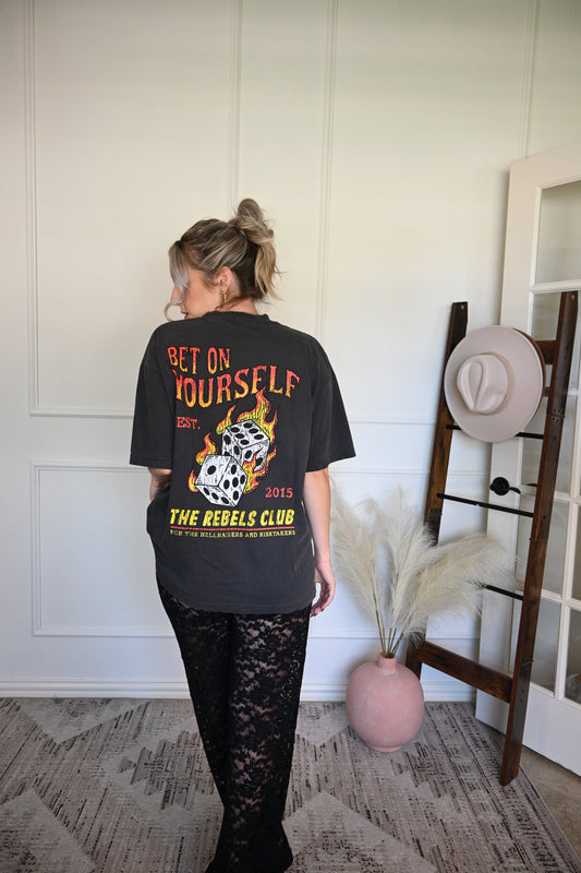 Person wearing a black graphic tee with text and graphics, standing in front of a wall.