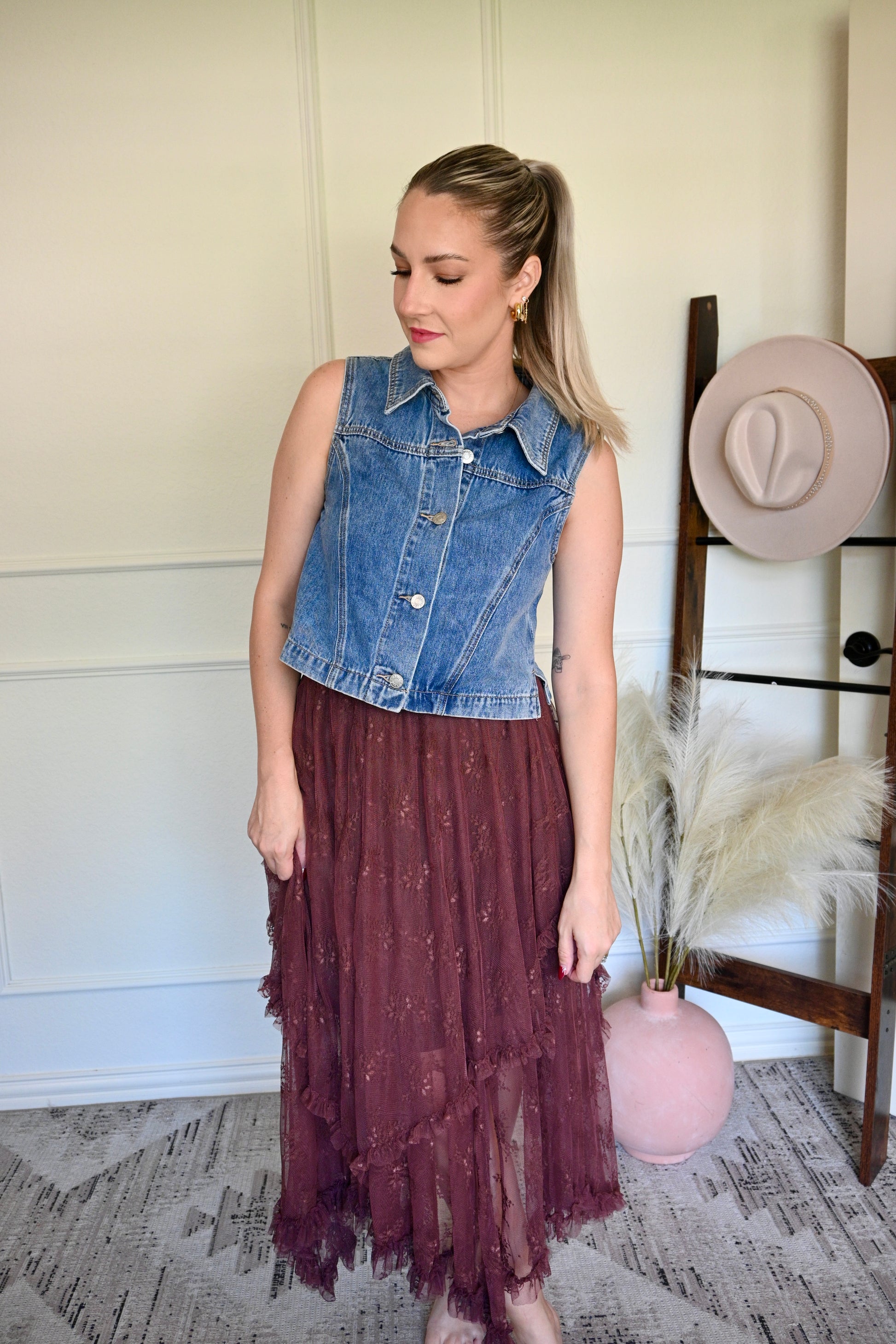 Woman wearing a denim vest with a brown lace skirt in a room with decorative elements.