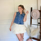 Woman in denim vest and white skirt standing in a room with a hat rack and vase.