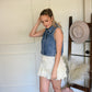 Woman wearing a denim vest with  ivory lace shorts and cowgirl boots standing next to a hat hung on a ladder.