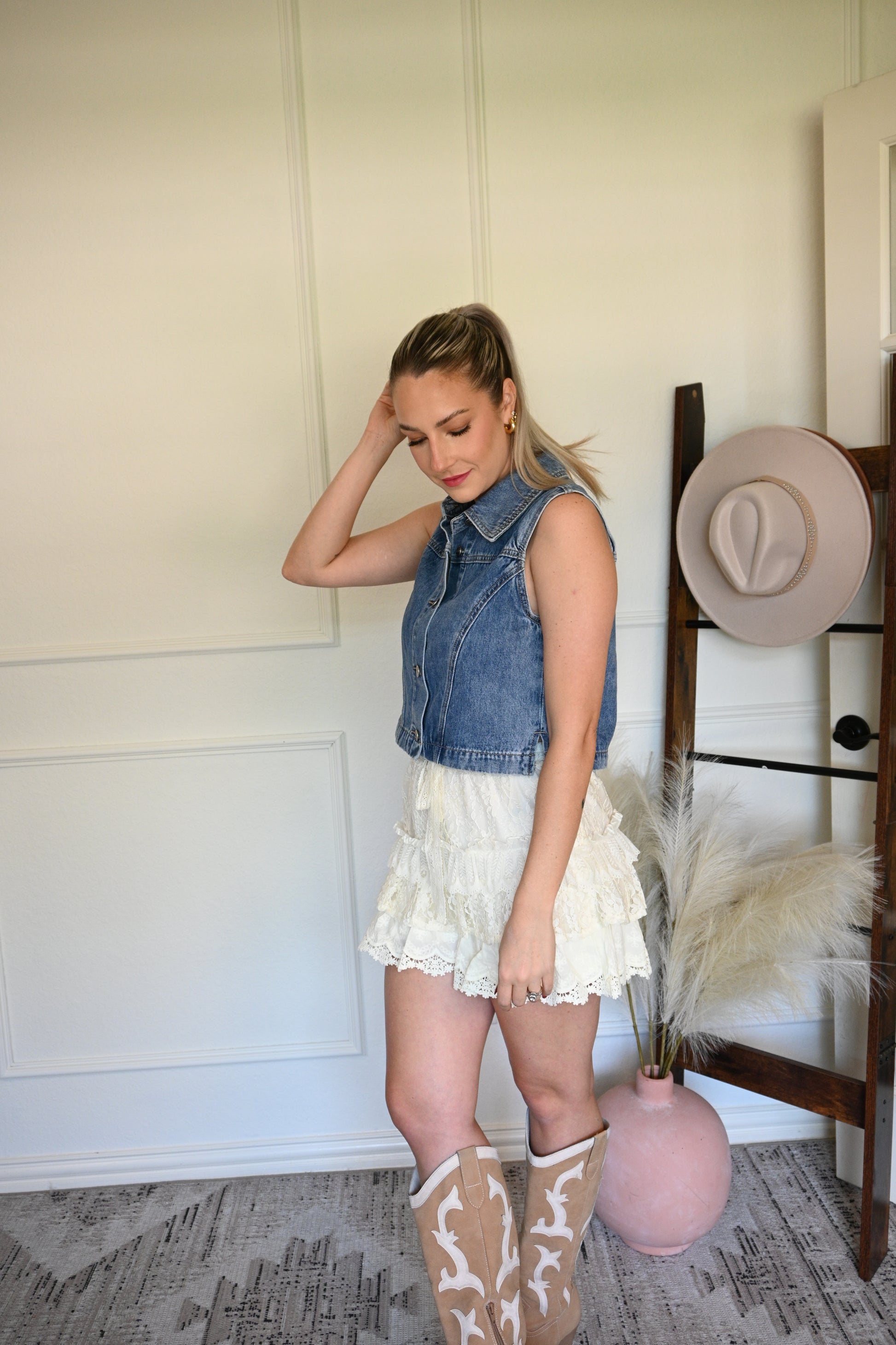 Woman wearing a denim vest with  ivory lace shorts and cowgirl boots standing next to a hat hung on a ladder.