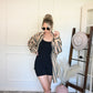Person wearing a black one piece and striped button down opened with a white wall and decorative elements in the background