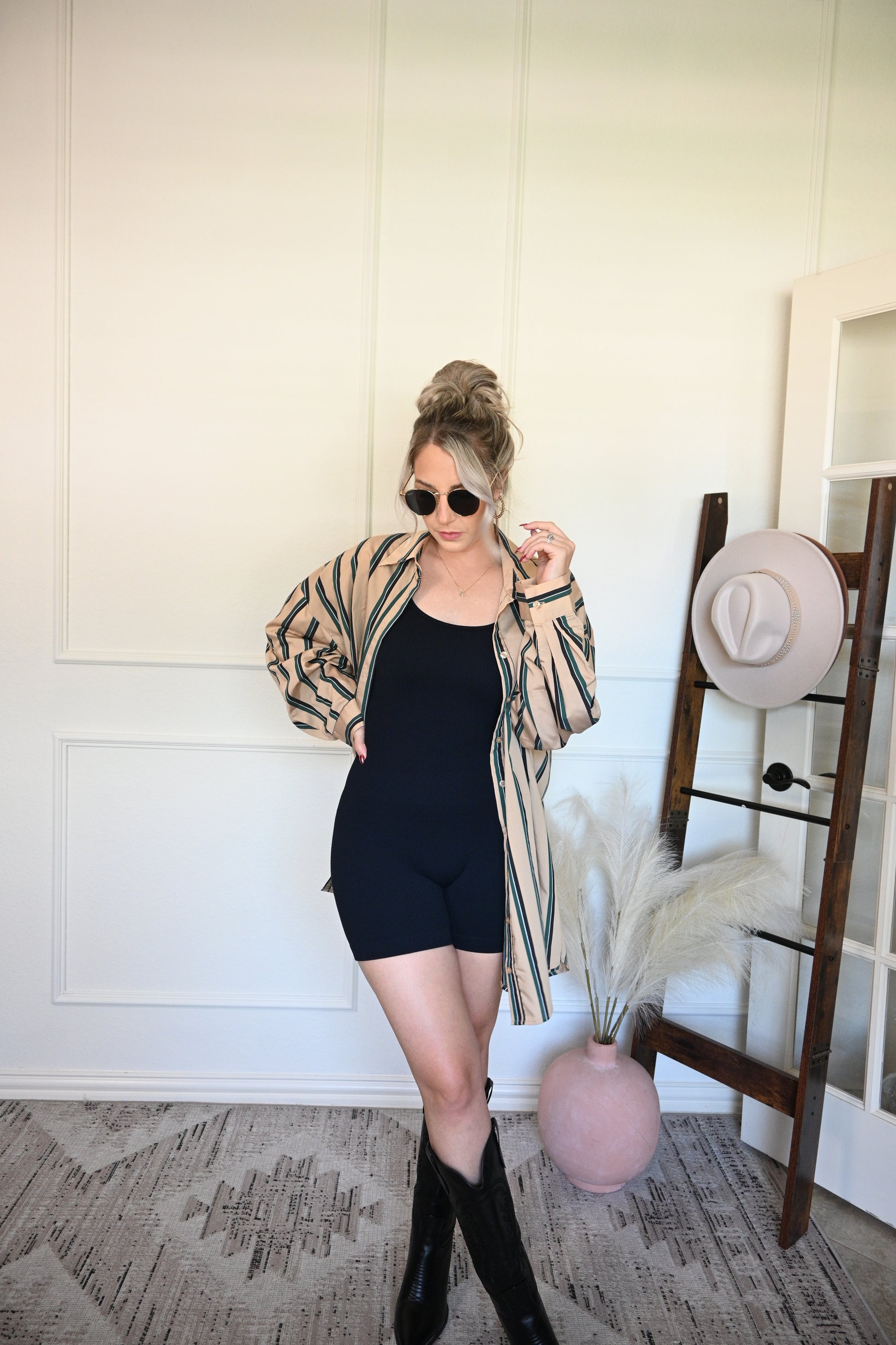 Person wearing a black one piece and striped button down opened with a white wall and decorative elements in the background
