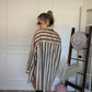 Woman in a striped shirt/dress standing next to a hat rack with hats on a white wall.