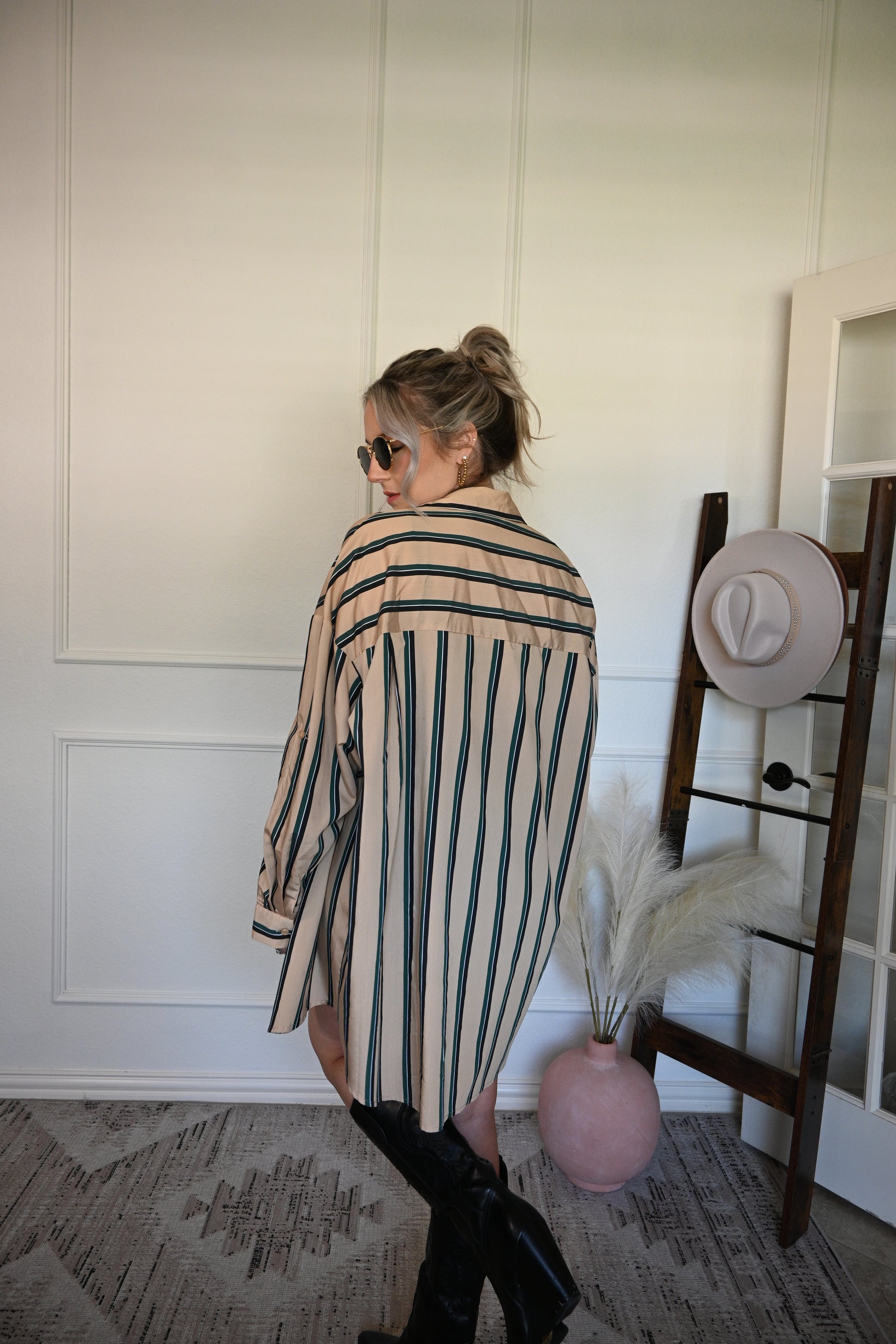 Woman in a striped shirt/dress standing next to a hat rack with hats on a white wall.