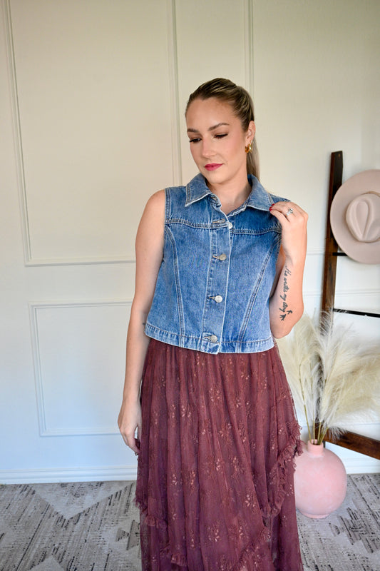 Woman wearing a denim vest and a brown skirt, standing indoors.