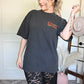 Woman wearing a washed black t-shirt with text, standing in a room with a white wall and decorative items
