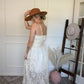 Woman in a ivory top and lace skirt set and brown hat standing next to a hat rack with hats on it.