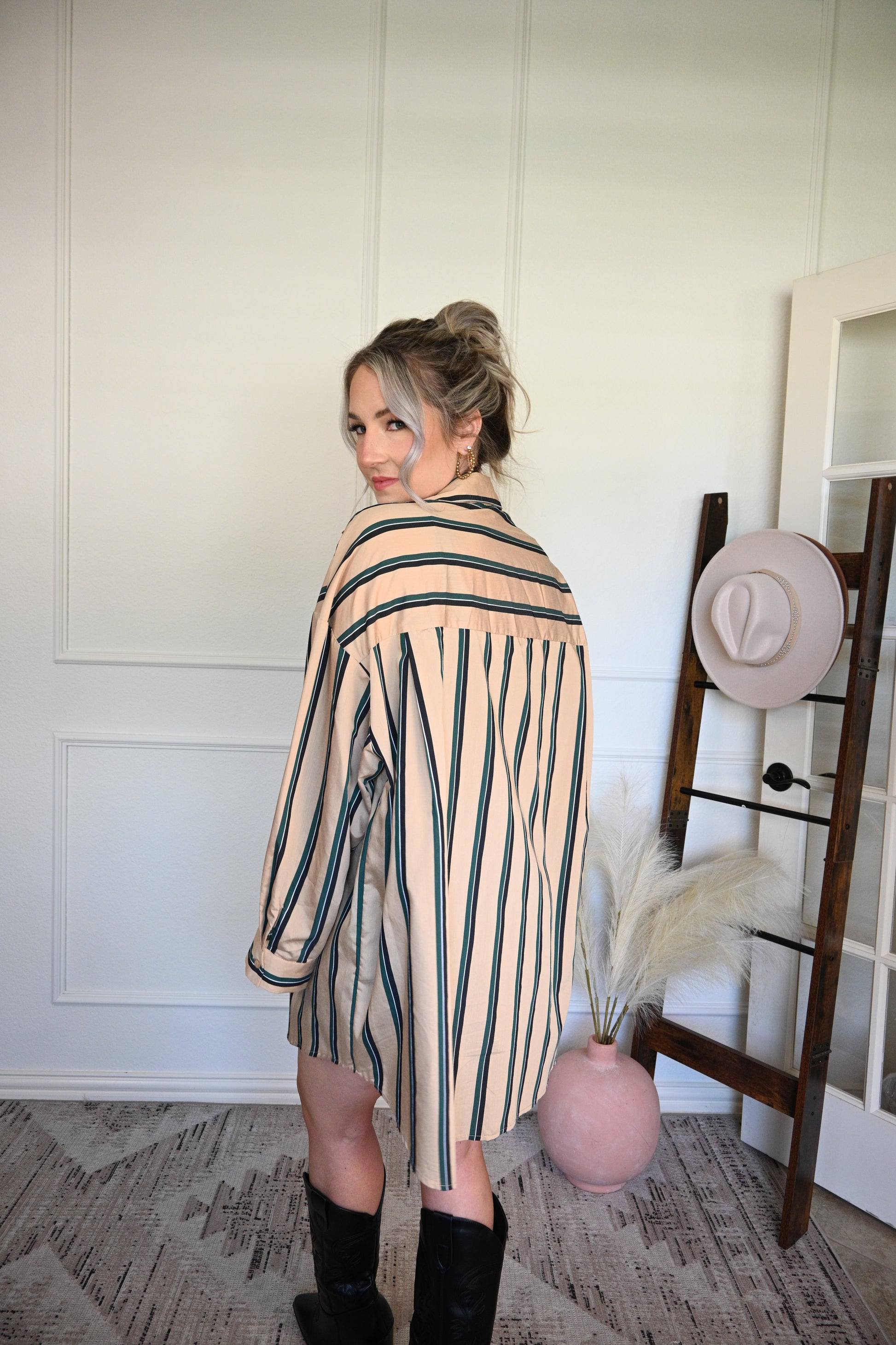 Woman wearing a striped shirt/dress standing next to a wooden stool with a hat on it against a white wall.