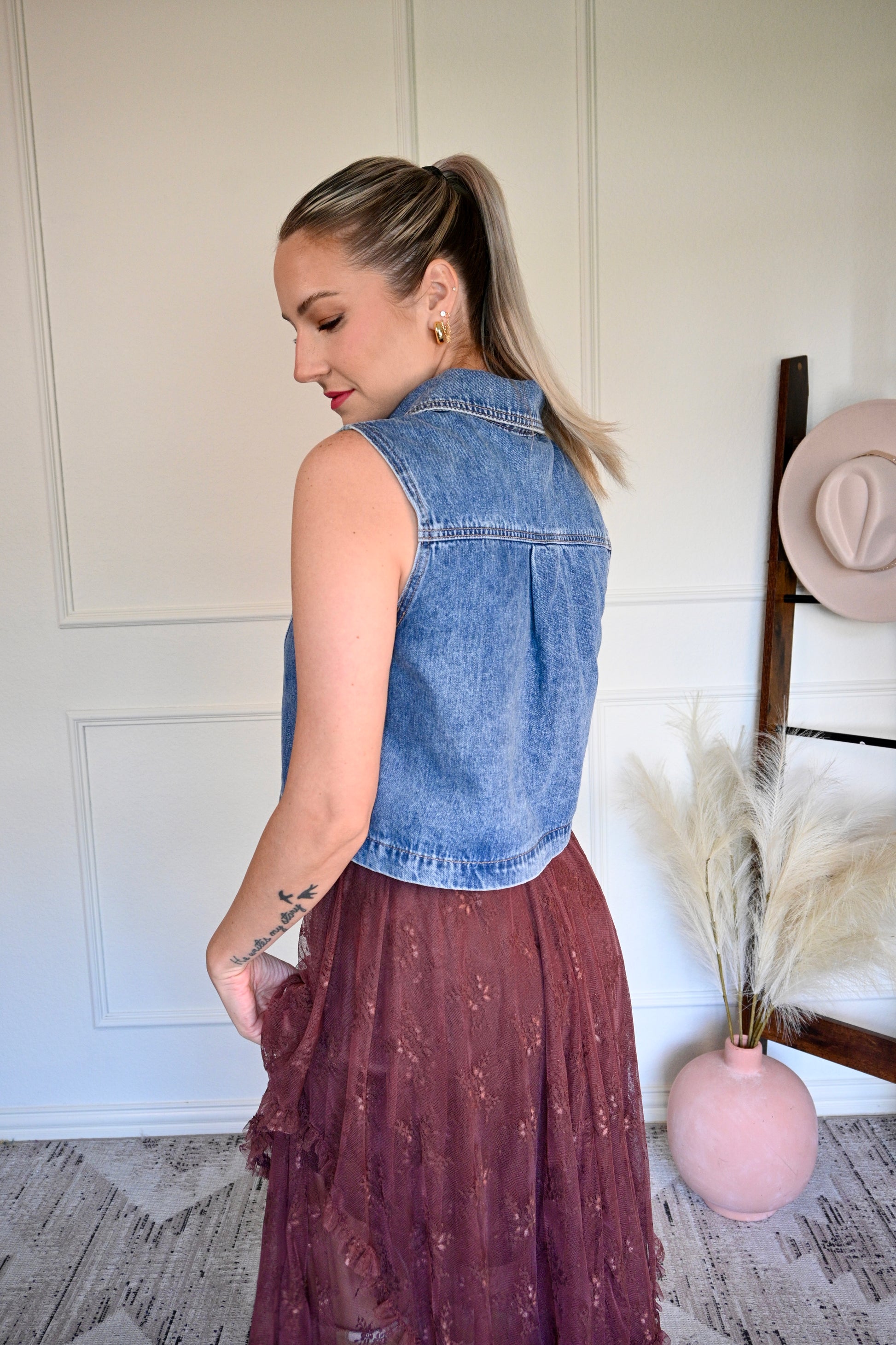 Woman wearing a denim vest with a brown lace skirt in a room with a pink vase and hat on a rack.