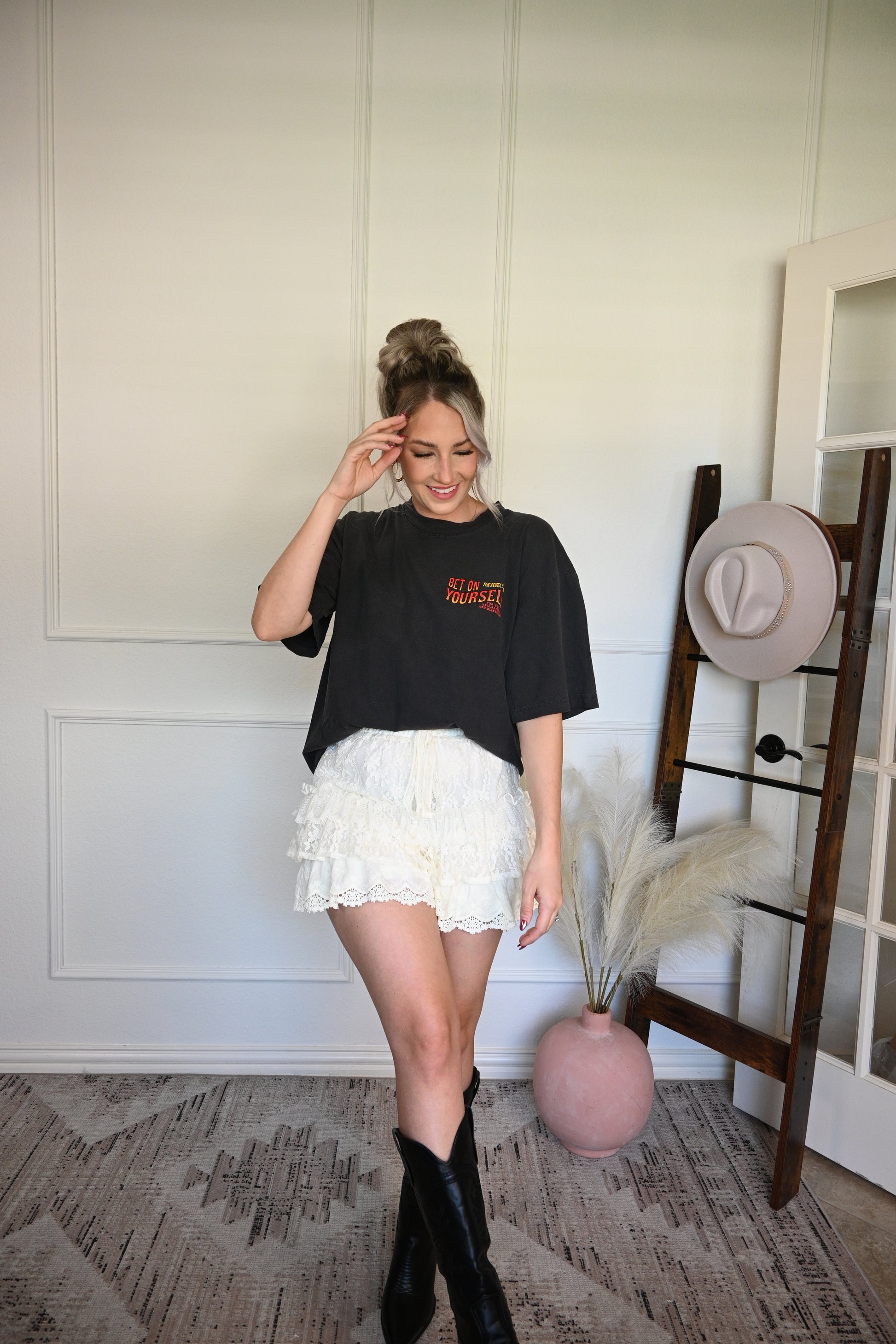 Person wearing a black graphic tee and cream ruffled skirt with black boots standing on a decorative rug.