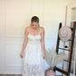 Woman in a ivory tank top and lace skirt set standing next to a hat on a stand in a room with a textured wall.